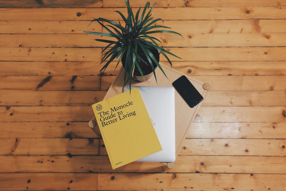 A top-down view of a small, square wooden table on a polished honey-colored wooden floor. The table holds a green potted plant with long, slender leaves, a closed laptop with a silver cover, a black s