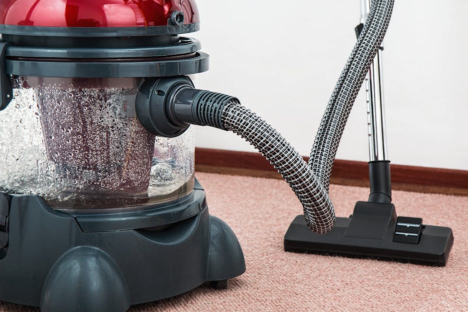A woman in casual clothing, including a beige jacket and jeans, is using a vacuum cleaner with a yellow body and black hose to deep clean a patterned area rug in a living room. The room features woode