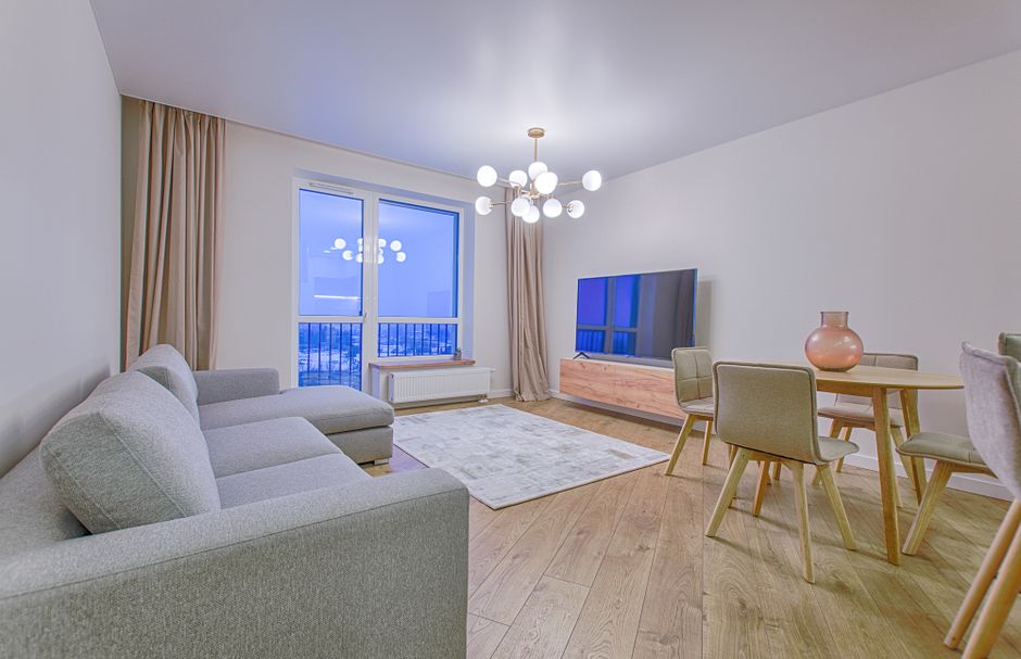 A spacious, well-lit living room featuring a light-colored fabric sofa and a matching armchair arranged around a white and beige area rug on a wooden floor. Large windows with beige curtains let in na