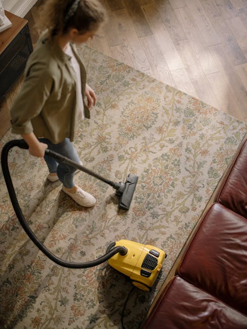A person kneeling on a patterned carpet in a well-lit room, operating a yellow and black portable carpet cleaning device. The individual is wearing a beige jacket and blue jeans, and is actively press
